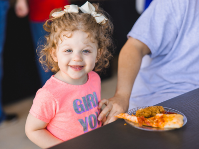 A young girl sits at a table with a plate of pizza and chicken nuggets in front of her.